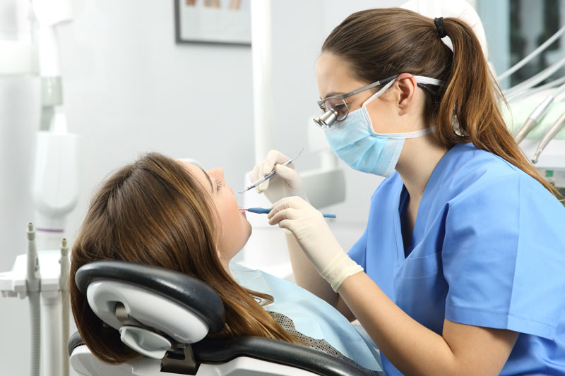 Dentist wearing eyeglasses gloves and mask examining a patient tooth with a dental probe and a mirror