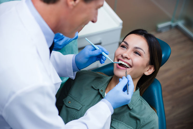 Female dental patient smiling during a checkup as a male dentist examines her teeth with dental tools in a modern dental office.